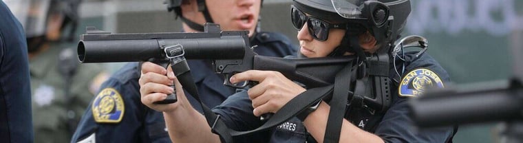 Police officer in tactical gear aiming a less-lethal weapon during a law enforcement operation. Proper training and deployment of less-lethal weapons ensure safe de-escalation and compliance.