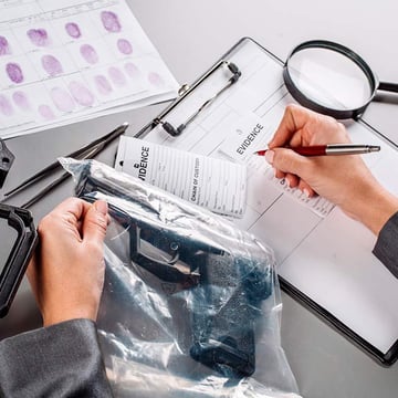 Evidence technician documenting chain of custody on an intake form beside a sealed evidence bag and fingerprint cards